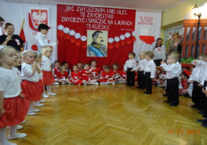Na tle biało czerwonej dekoracji stoją naprzeciwko siebie odświętnie ubrane dzieci.